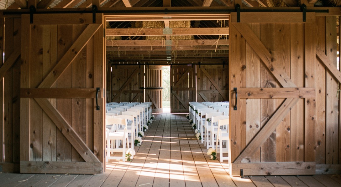Open barn doors framing a ceremony aisle of white chairs, hayloft beams overhead and late afternoon sun cutting across the plank floor
